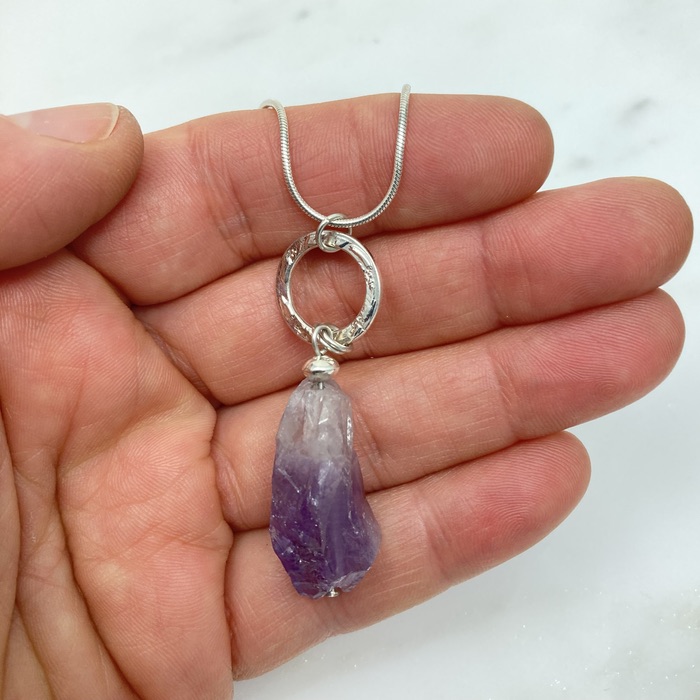 Amethyst rough cut nugget pendant with patterned sterling ring on a sterling snake chain. It's held in a white person's hand.