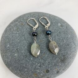 Faceted oval Labradorite and deep purple-blue freshwater pearl earrings on sterling leverback ear wires displayed on a round grey beach rock.