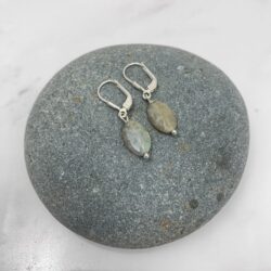 Faceted oval Labradorite earrings on sterling ear wires. The stone is translucent pale olive green, with iridescent facets when the light hits them. The earrings are displayed on a small round grey beach rock.