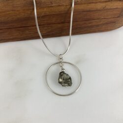 Multifaceted pyrite nugget sits within a sterling circle, hanging on a sterling snake chain. The pendant sits on white marble, the chain draped over a small carved wooden box.