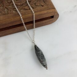 Closeup of a faceted elongated oval black and cream silver leaf jasper pendant on a sterling chain. The chain is draped over a small carved wooden box. The pendant is sitting on white marble.